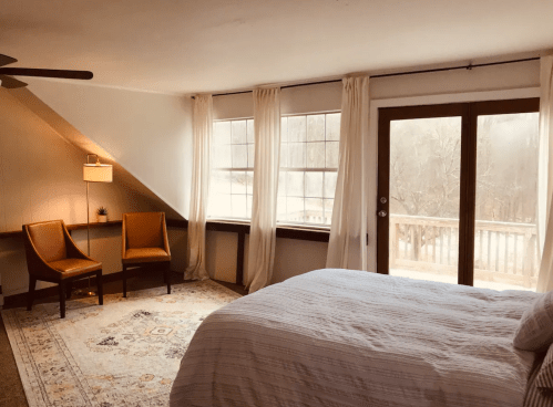 Cozy bedroom with a bed, two chairs, large windows, and a door leading to a deck, featuring soft lighting and a rug.