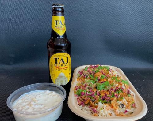 A plate of colorful biryani topped with herbs, a side of yogurt, and a bottle of Taj Mahal beer.