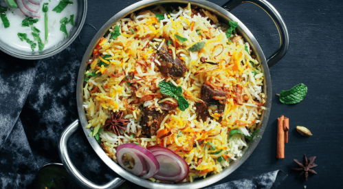 A pot of colorful biryani with spices, garnished with mint and onions, alongside a bowl of yogurt.