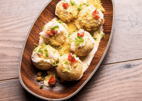 A wooden platter with six crispy snacks topped with yogurt, tomatoes, and herbs, served on a wooden table.
