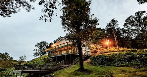 A modern house on a hillside, surrounded by trees, with warm lights glowing at dusk.
