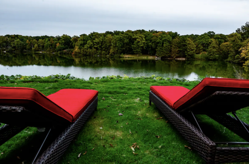 Two red lounge chairs on green grass overlooking a calm lake surrounded by trees under a cloudy sky.
