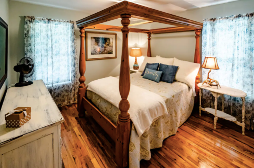 Cozy bedroom with a four-poster bed, decorative pillows, wooden furniture, and natural light from two windows.