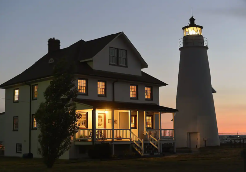 A lighthouse beside a two-story house, both illuminated at dusk, with a clear sky in the background.