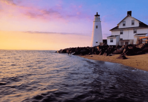 A lighthouse beside a rocky shore at sunset, with calm waters reflecting the colorful sky.