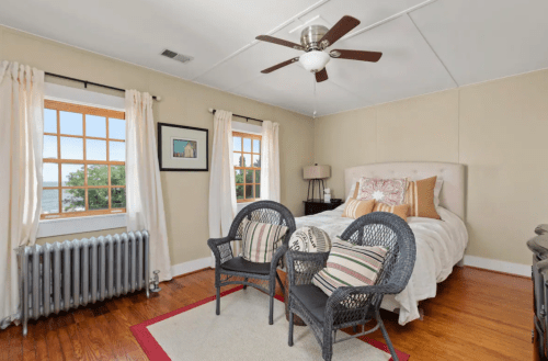 Cozy bedroom with a bed, two chairs, large windows, and a radiator, featuring light-colored walls and wooden flooring.