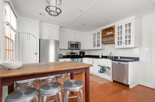 Bright kitchen with white cabinets, stainless steel appliances, a wooden table, and bar stools. Natural light from windows.