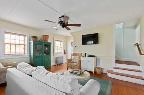 Cozy living room with a white sofa, green cabinet, TV, and wooden stairs leading to an upper level. Bright and inviting space.
