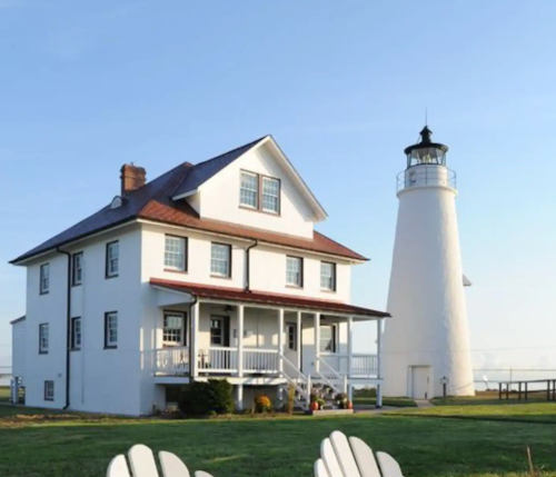 A white house with a porch next to a tall lighthouse against a clear blue sky. Green grass in the foreground.