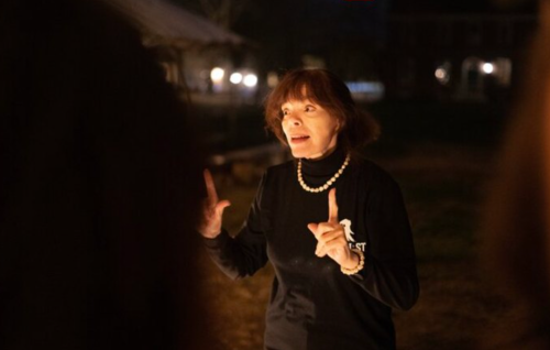 A woman in a black shirt gestures while speaking passionately in a dimly lit outdoor setting.