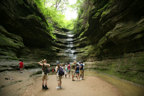 A group of people exploring a lush, rocky canyon with a waterfall in the background.