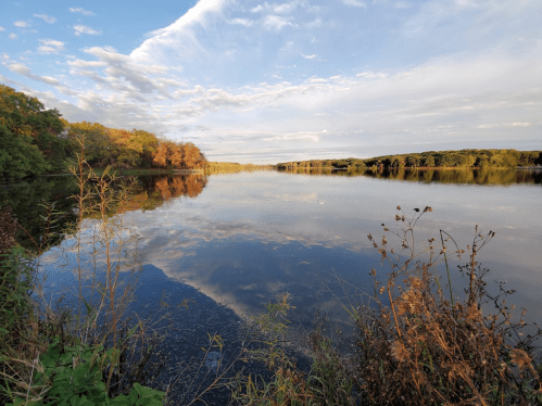 A serene river scene with reflections of clouds and trees, surrounded by lush greenery and autumn foliage.