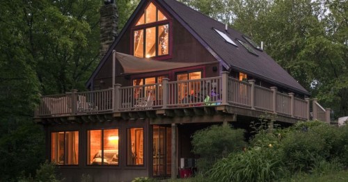 A cozy, modern cabin with large windows and a deck, surrounded by lush greenery at dusk.