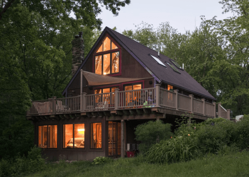 A cozy, modern house with large windows, surrounded by greenery, illuminated at dusk.