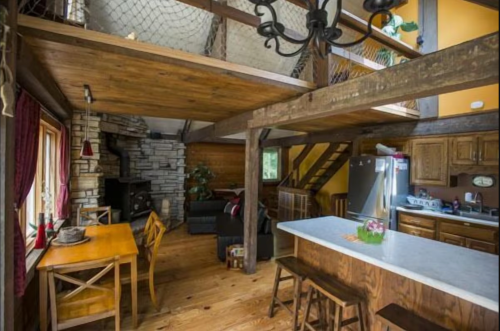 Cozy wooden cabin interior with a dining area, kitchen, and living space featuring a stone fireplace and rustic decor.