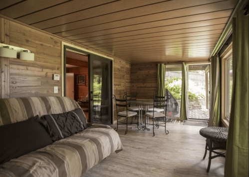 Cozy interior of a wooden cabin featuring a sofa, dining table, and large windows with greenery outside.