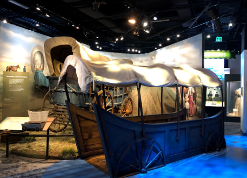 A covered wagon displayed in a museum, with historical context and other artifacts in the background.