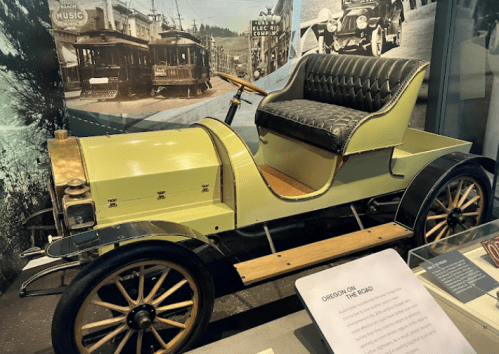 A vintage car with a green body and black leather seat displayed in a museum, surrounded by historical images.
