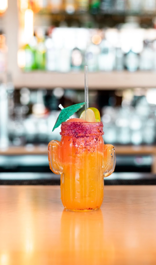A colorful cocktail in a cactus-shaped glass, garnished with a lime and a small umbrella, on a wooden bar.
