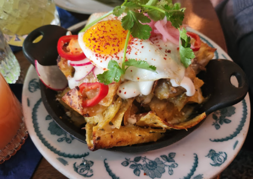 A skillet of nachos topped with a fried egg, sour cream, pickled onions, and fresh herbs, served on a decorative plate.