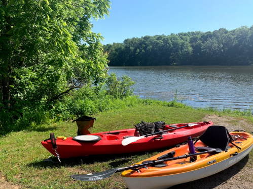 Two kayaks, one red and one orange, are parked on the grassy shore by a calm lake surrounded by trees.