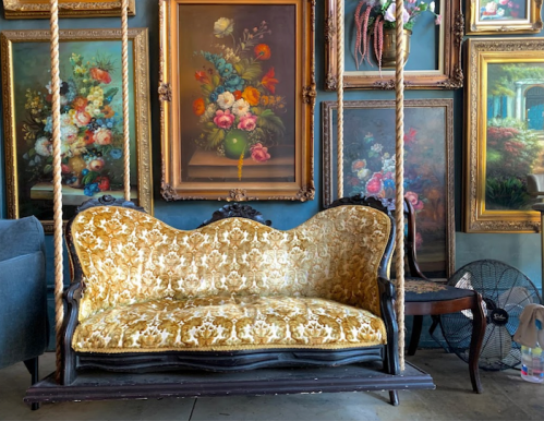 A vintage swing sofa with a floral cushion, surrounded by ornate framed paintings on a wall.