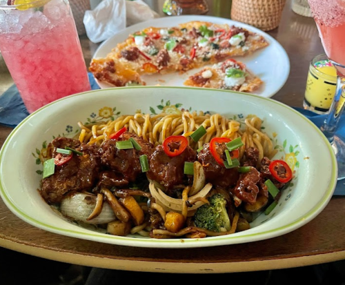 A bowl of noodles with beef, vegetables, and chili, with a pizza and drinks in the background.