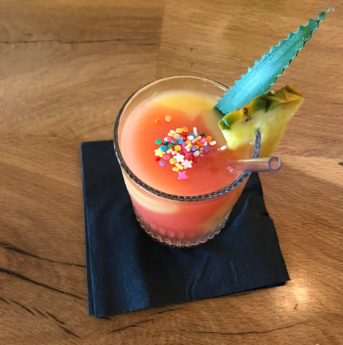 A colorful cocktail with sprinkles and a pineapple slice, served in a glass on a black napkin.