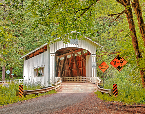 A white covered bridge surrounded by greenery, with signs indicating a one-lane bridge and height restriction.