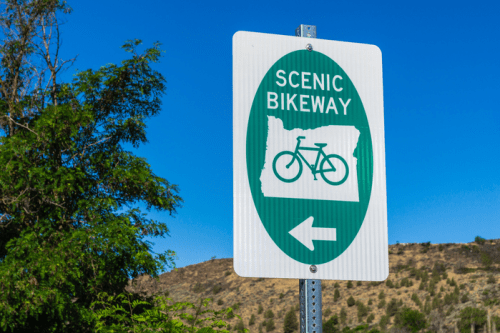 Sign for a scenic bikeway with a bicycle icon, pointing left against a clear blue sky and green foliage.