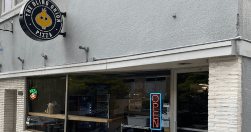 Exterior of The Blind Onion Pizza, featuring a large window, "OPEN" sign, and outdoor seating.