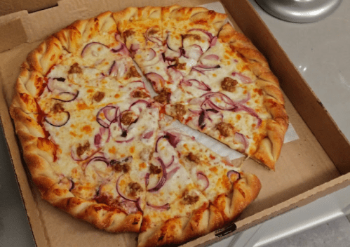 A freshly baked pizza with cheese, red onions, and sausage, cut into slices, in a cardboard box.
