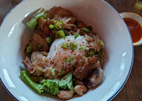 A bowl of grilled meat with lettuce, topped with a poached egg, sliced green chilies, and chili flakes, served with sauce.