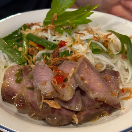 A close-up of a dish featuring sliced meat, rice noodles, herbs, and crispy onions, garnished with chili and greens.