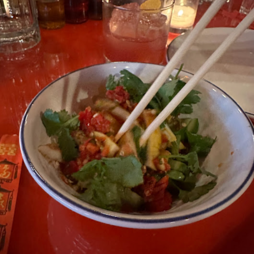 A bowl of salad with fresh herbs and vegetables, chopsticks resting on the edge, on a vibrant orange table.