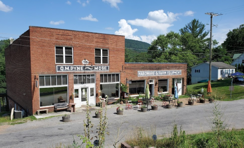 A brick building with "Q.M. Dyne" sign, featuring a hardware and farm equipment store, surrounded by potted plants.