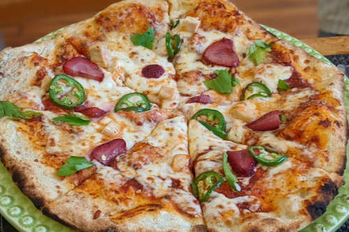 A freshly baked pizza topped with cheese, jalapeños, chicken, strawberries, and fresh herbs on a green plate.