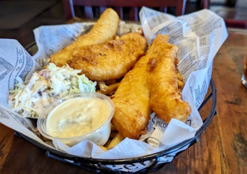 A basket of golden fried fish fillets with a side of coleslaw and dipping sauce on a wooden table.