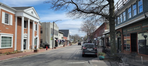A quiet street lined with brick buildings, shops, and parked cars under a clear blue sky.