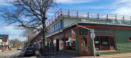 A street view of a colorful building with a rooftop deck, featuring a pub sign, surrounded by trees and shops.