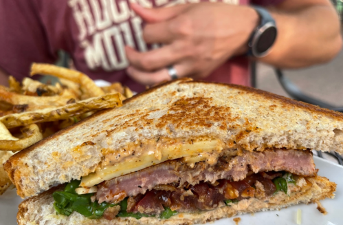 A close-up of a sandwich with layers of meat, cheese, and greens, served with a side of fries.