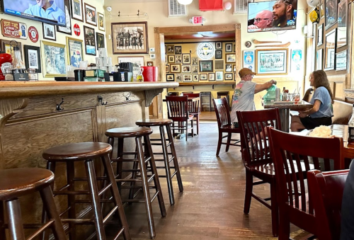 A cozy bar with wooden stools, framed photos on the walls, and two patrons engaged in conversation at a table.