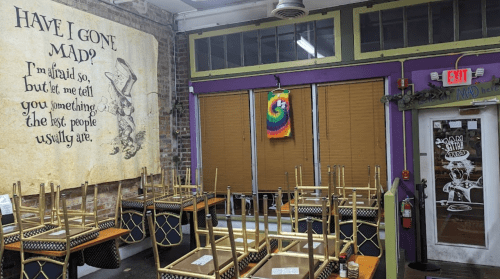 A colorful restaurant interior with stacked chairs, a vibrant wall art, and a sign reading "Have I gone mad?"