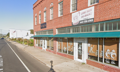 A brick building with large windows and green awnings, featuring a sign that invites customers to "Come Taste the Magic!"