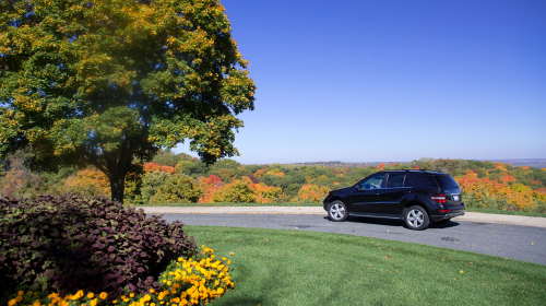 A black SUV parked near a scenic overlook with vibrant autumn foliage and a clear blue sky.