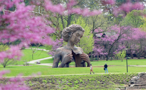 A large statue of a woman stands in a park surrounded by blooming pink trees, with two people walking nearby.