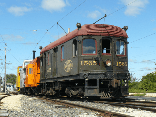 A vintage train with a black and red locomotive and an orange car, traveling along a railway under a blue sky.
