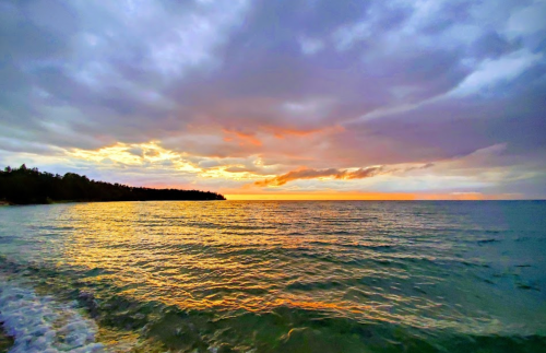 A serene beach scene at sunset, with colorful clouds reflecting on calm waters and a distant shoreline.