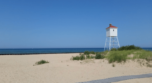 A white lighthouse on a sandy beach with green grass and a clear blue sky above the calm ocean.