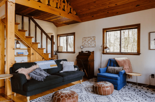 Cozy living room with a black sofa, blue armchair, wooden stairs, and large windows overlooking a forested area.